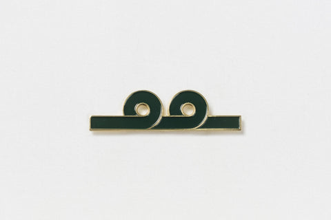 A rectangular green pin with gold edges and two upward loops in the design in the center